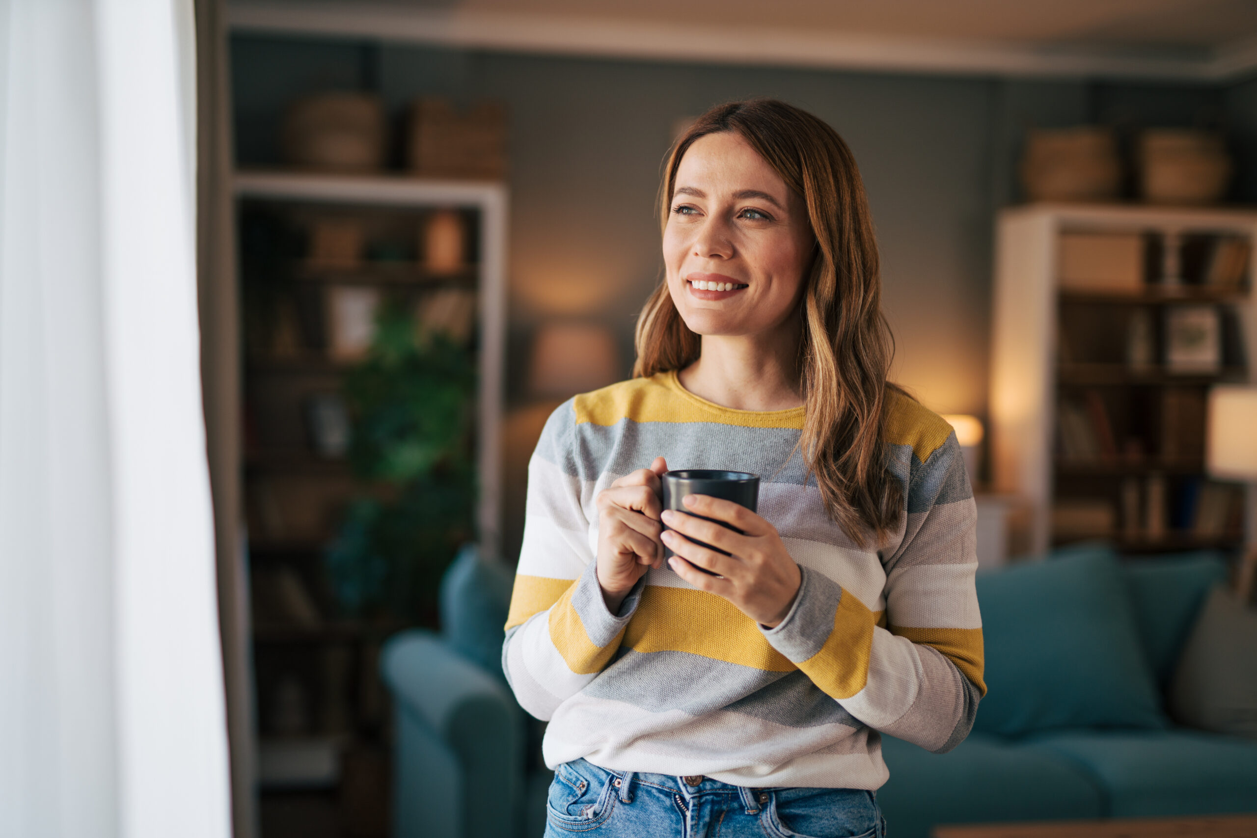 Portrait of a mature woman looking out the window in her living room and drinking her morning coffee.