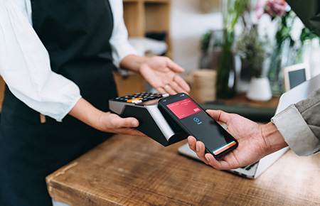 Close up of young man shopping at the flower shop. He is paying with his smartphone, scan and pay a bill on a card machine making a quick and easy contactless payment.