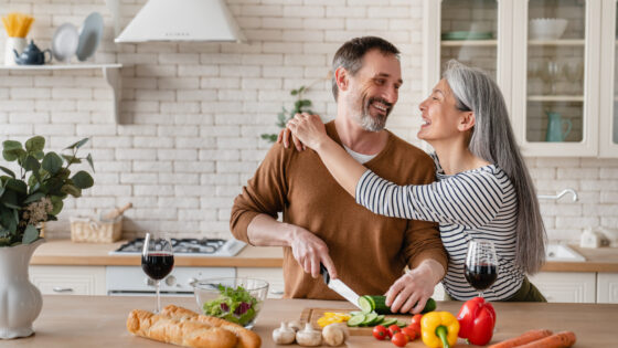 Cheerful mature husband man cutting vegetables cooking food meal in the kitchen while his wife woman embracing hugging him helping prepare salad together at home.