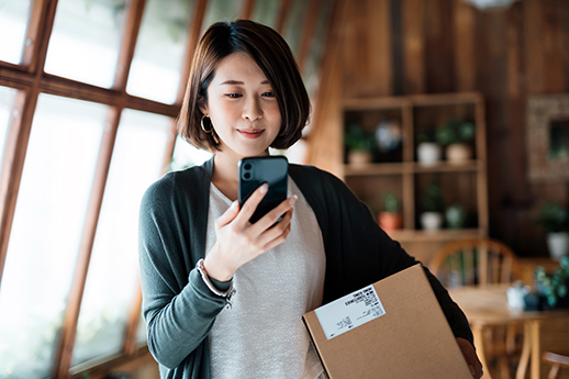 Smiling young Asian woman shopping online with smartphone on hand, receiving a delivered parcel by home delivery service. Online shopping, online banking. Enjoyable customer shopping experience.