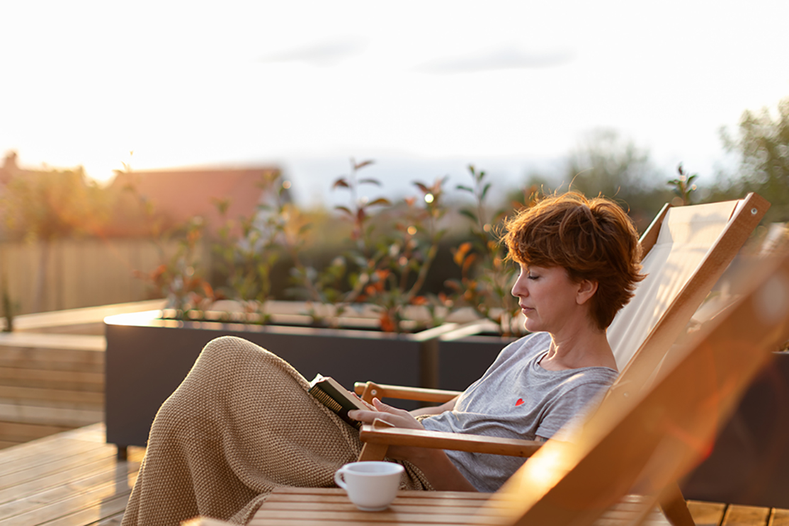 Beautiful woman is sitting on patio on the deck chair and enjoying beautiful surrounding while drinking coffee or tea and reading a book